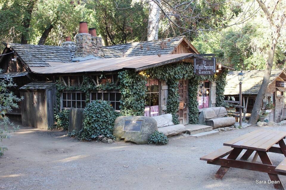 A rustic cabin with a sign reading "The Cold Spring Tavern" surrounded by trees and a wooden fence.