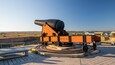 Un grand canon est installé sur une plateforme circulaire, adossé à un mur de briques et précédé d’un banc. L’ensemble se détache sur un ciel dégagé et des bâtiments situés au loin.