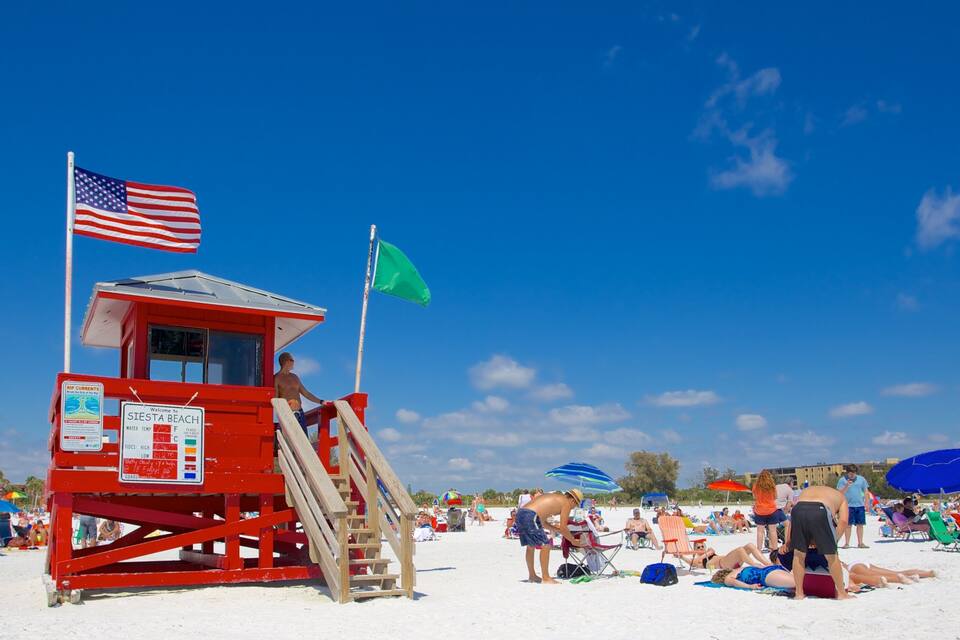 A lifeguard tower with an American flag, a green flag, and a signboard on a beach with people sunbathing and umbrellas.