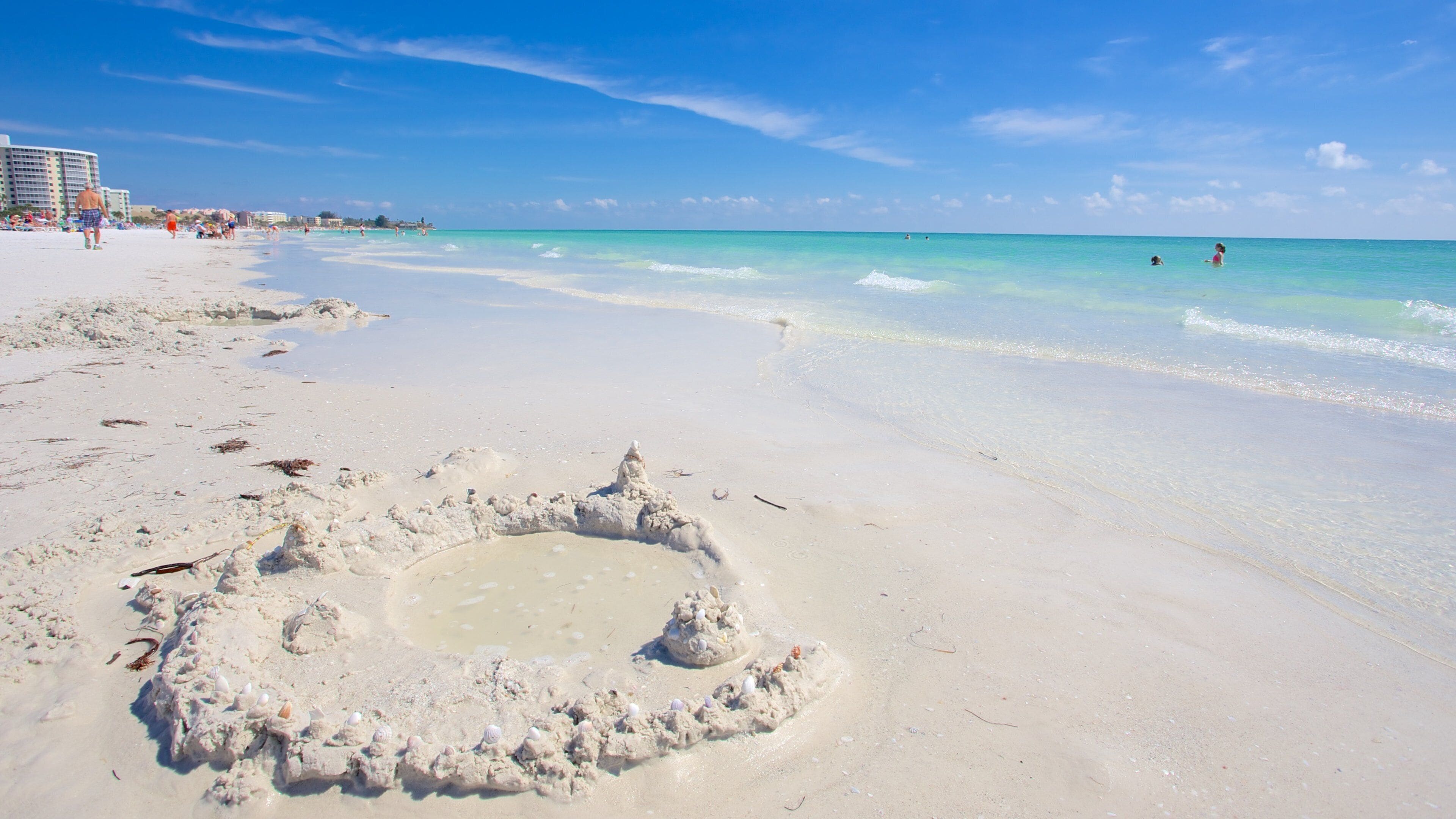 A sandy beach with clear turquoise water, a few people in the distance, and a sandcastle in the foreground.