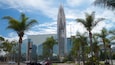 Een modern hoogbouwwerk met een spiegelende glazen gevel, omgeven door palmbomen en ander groen.