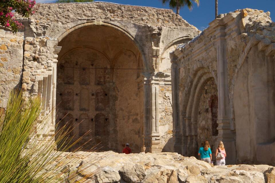 An ancient stone structure with arched doorways and columns, surrounded by rocky terrain and flowering plants.