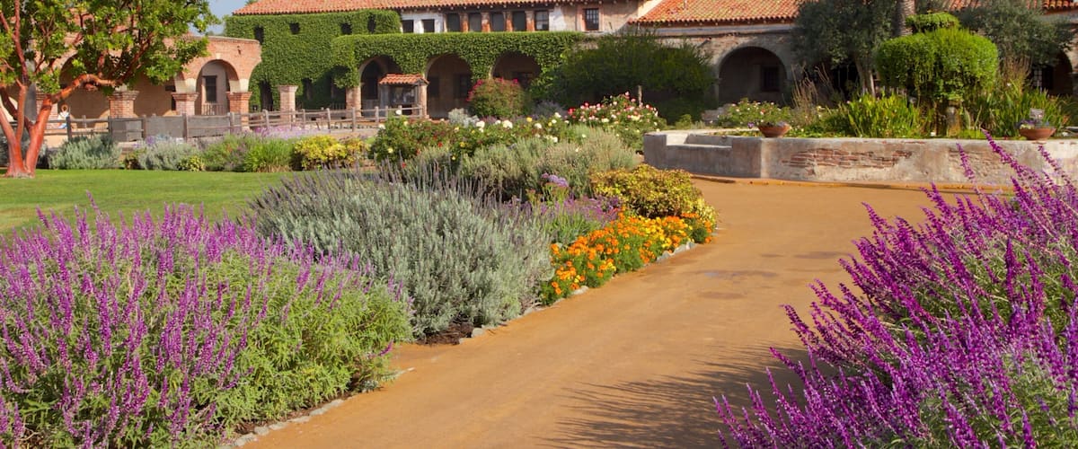 A historic building with red domes and arches, surrounded by a garden with purple flowers and a dirt path.