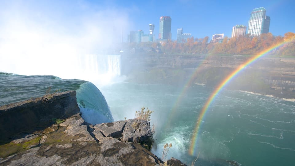 A waterfall with a rainbow, rocky terrain, and a city skyline in the background.