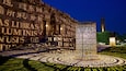 A large illuminated sculpture with inscriptions, surrounded by a patterned lawn and a building with classical architecture in the background.