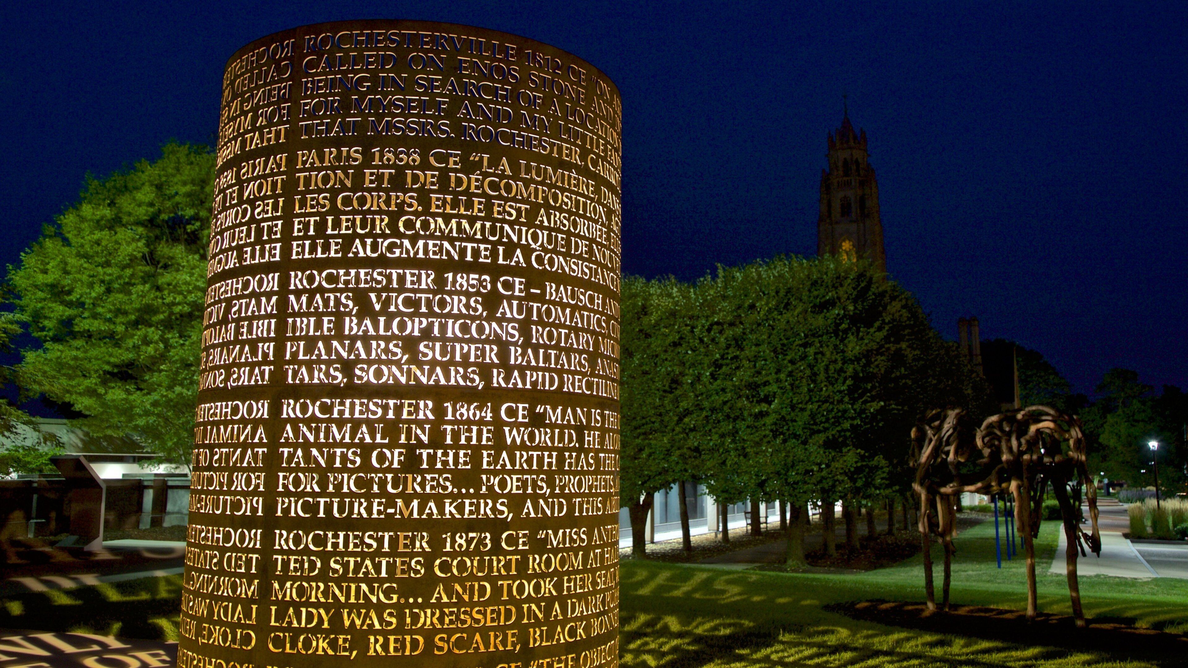 A cylindrical illuminated structure with historical text, set against a night sky with a church tower in the background.