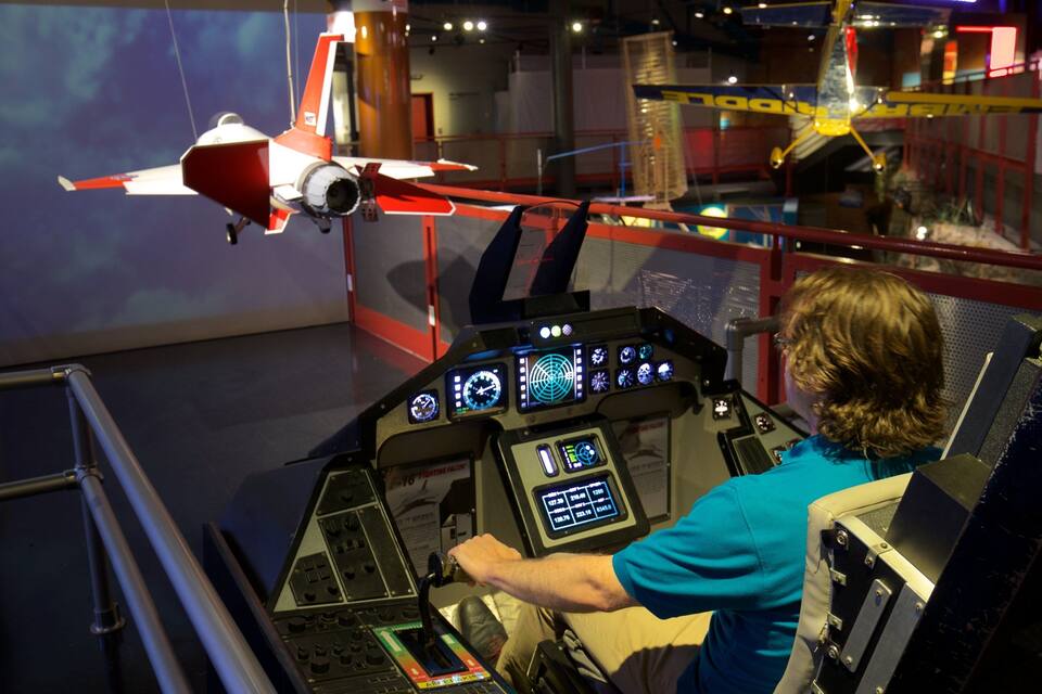 A person in a flight simulator cockpit with a model airplane in the background.