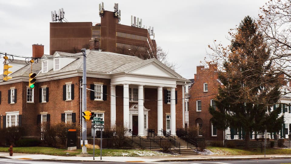 A historic building with a central portico, brick facade, and a large tree in the foreground.
