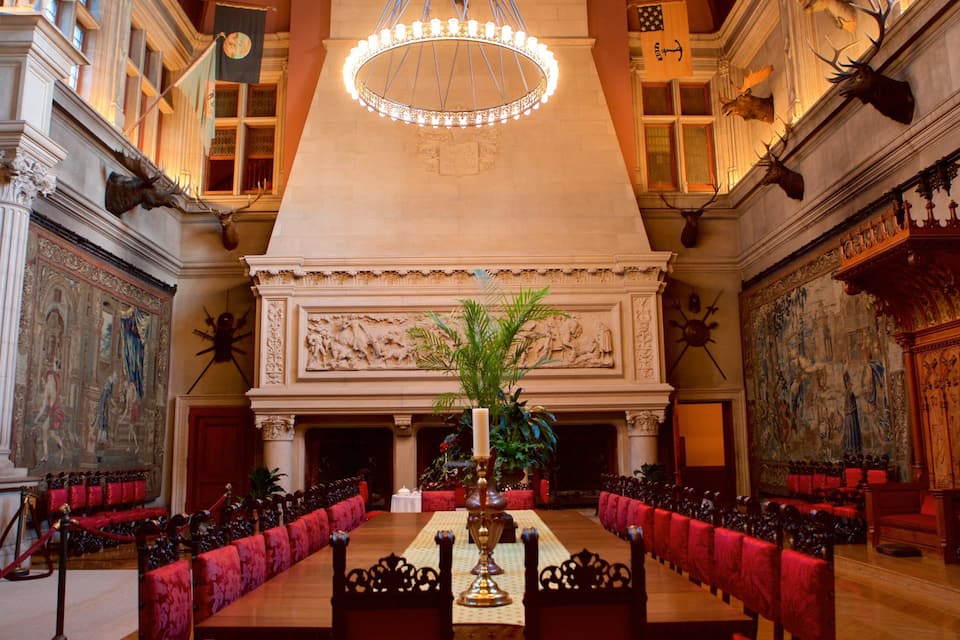 Un gran salón con techo alto, candelabros ornamentales y una larga mesa con sillas tapizadas en rojo.