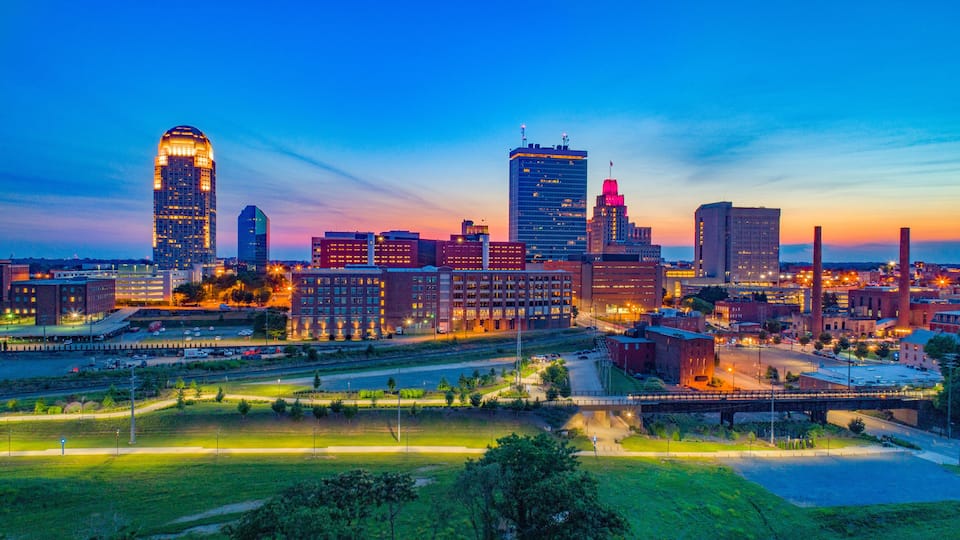 A city skyline at dusk with illuminated buildings and a bridge.