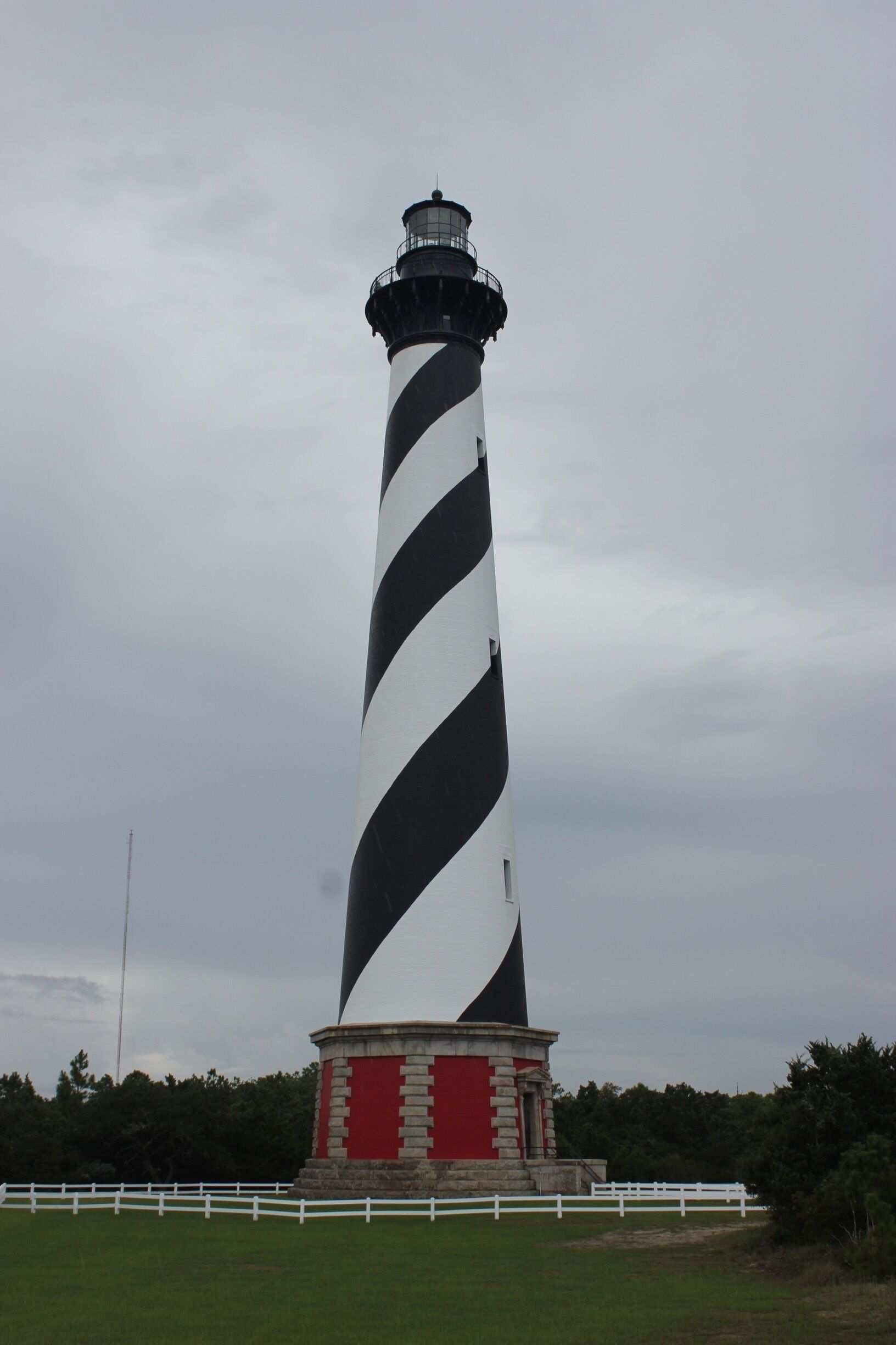 A tall lighthouse with black and white stripes, a red base, and a white fence in the foreground.