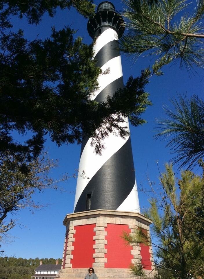 A black and white striped lighthouse with a red base, surrounded by pine trees.