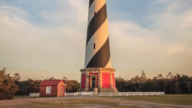 Um farol alto e listrado, com base vermelha, cercado por uma cerca branca e um pequeno edifício vermelho.