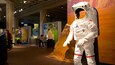En astronautdocka i naturlig storlek, klädd i en vit rymddräkt med röda ränder, som står framför en stor, bergig vägg i orange färg.