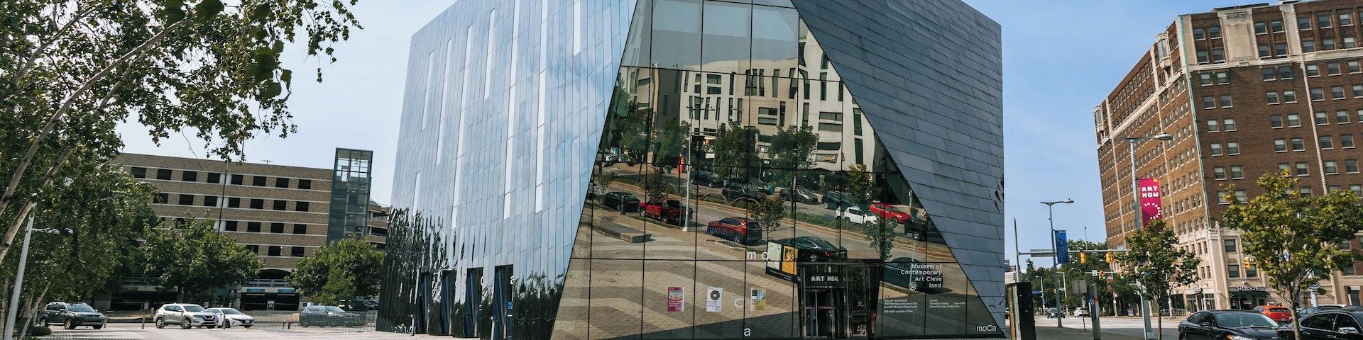 Um edifício moderno de vidro com fachada triangular, rodeado por ruas urbanas e outros edifícios.