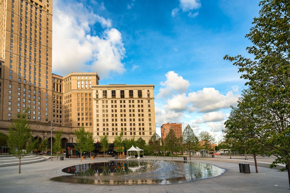 Una plaza urbana con una fuente, edificios altos y árboles.