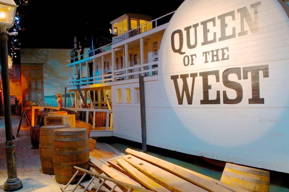 A model of the Queen of the West ship, surrounded by wooden barrels and a street lamp.