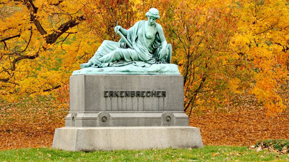Eine Statue einer Frau mit Kind in einem Park mit Herbstlaub.