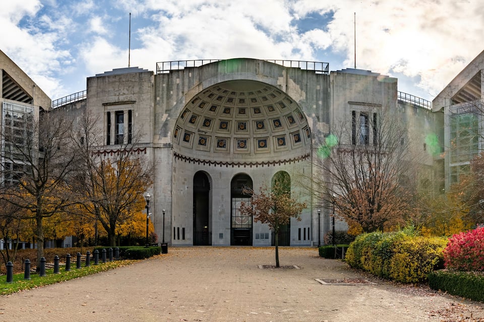 Un grande edificio storico con una cupola centrale e un ingresso ad arco, circondato da foglie autunnali e un vialetto lastricato.