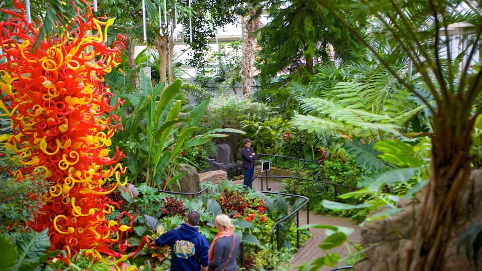 A lush indoor garden with vibrant glass sculptures, a variety of tropical plants, and a metal staircase.