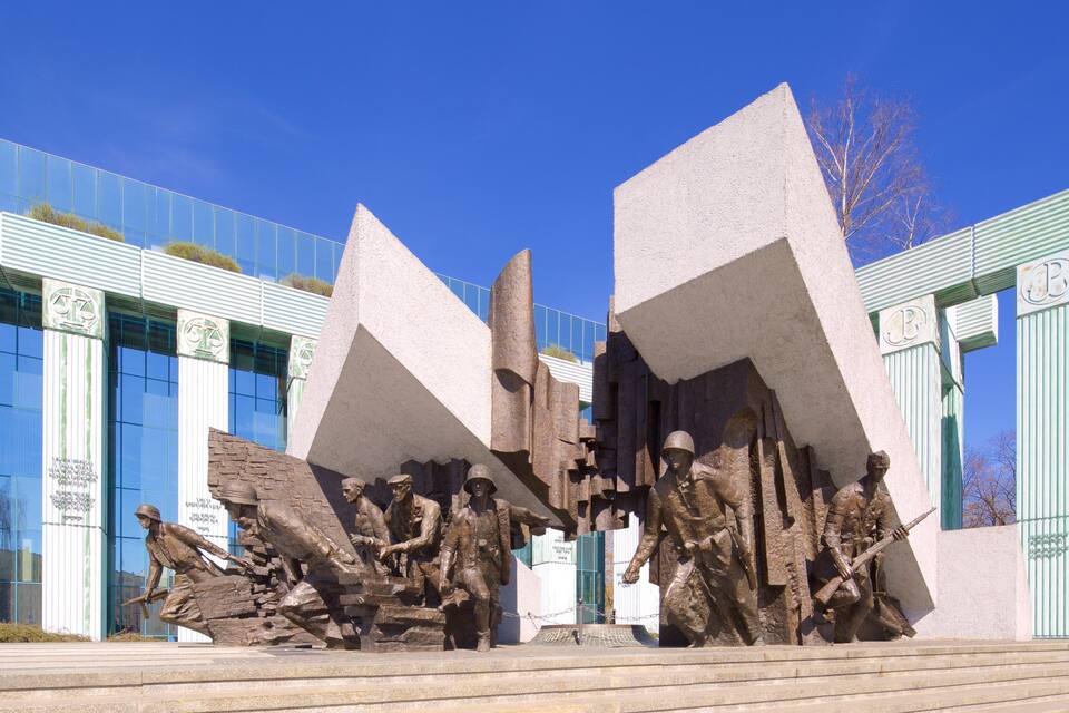 A bronze statue of soldiers in combat, set against a modern building with glass windows and green metal railings.
