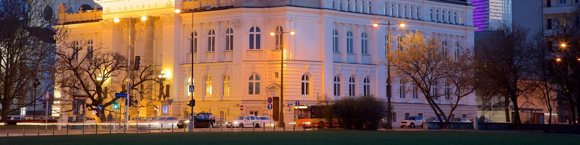 A historic building with a dome and a clock tower, surrounded by modern buildings and a park.