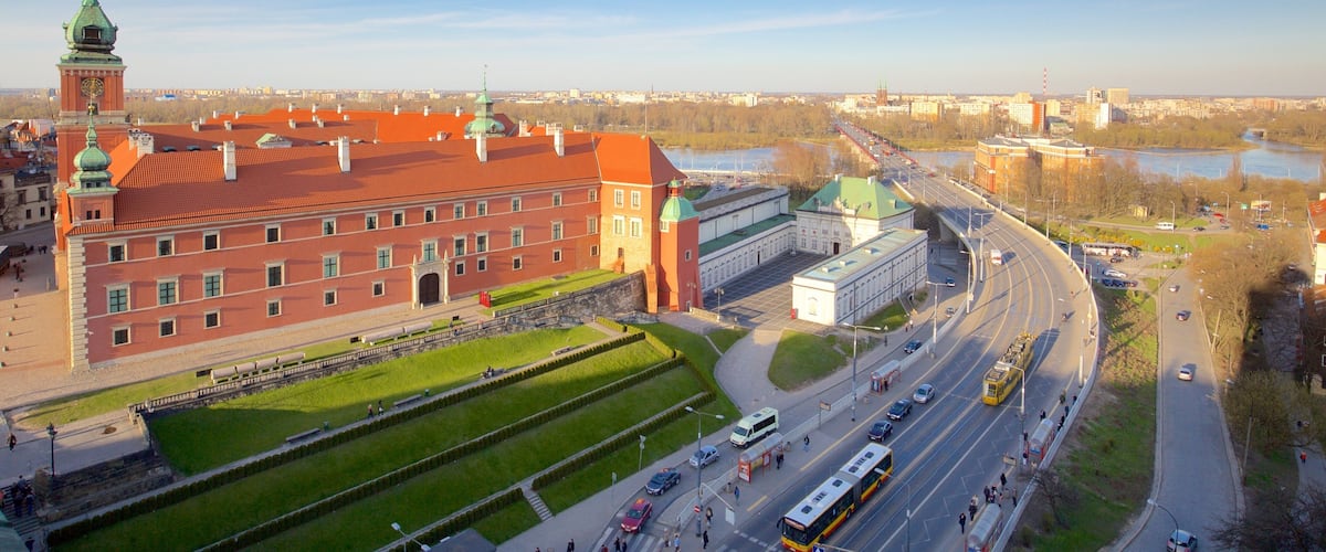 A large red brick building with a green dome and a clock tower, surrounded by green lawns and a river.