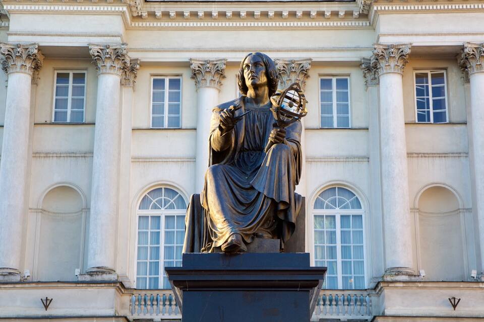 Una estatua de una figura sentada sosteniendo un globo terráqueo, frente a un edificio clásico con columnas y ventanas arqueadas.