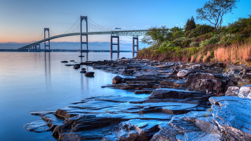 A suspension bridge over calm water, rocky shoreline, and dense greenery.