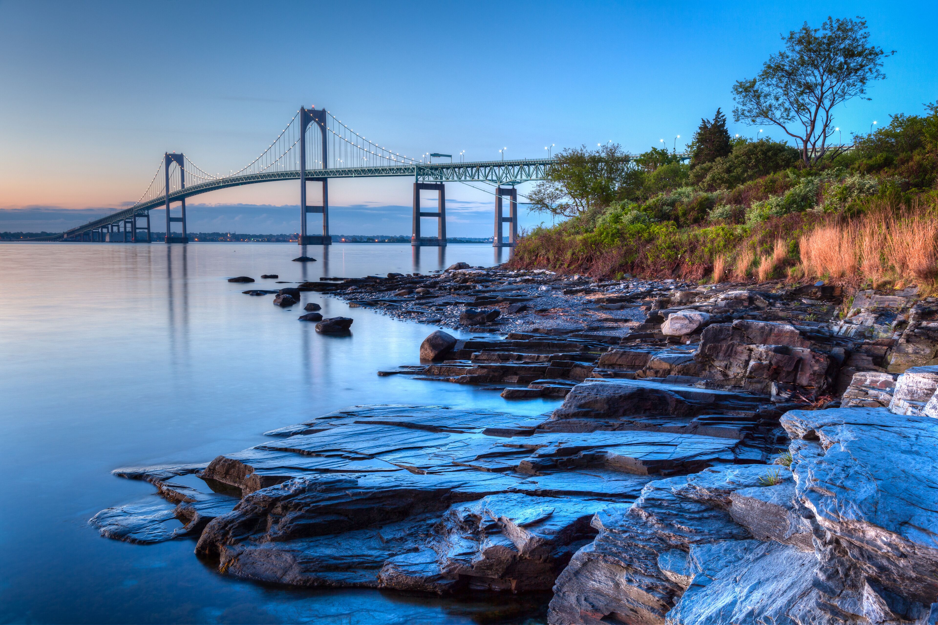 A suspension bridge over calm water, rocky shoreline, and dense greenery.