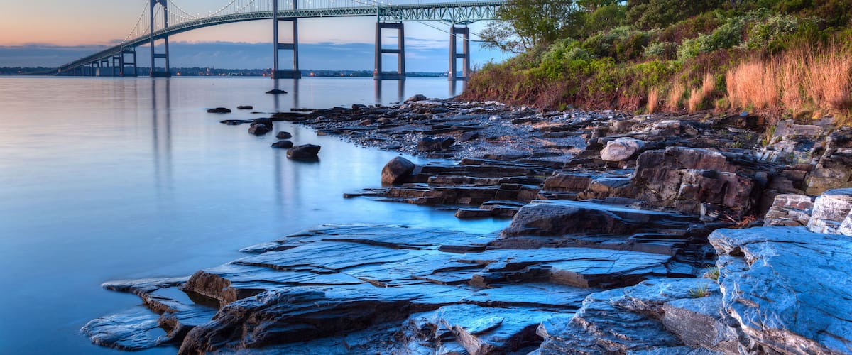 A suspension bridge over calm water, rocky shoreline, and dense greenery.