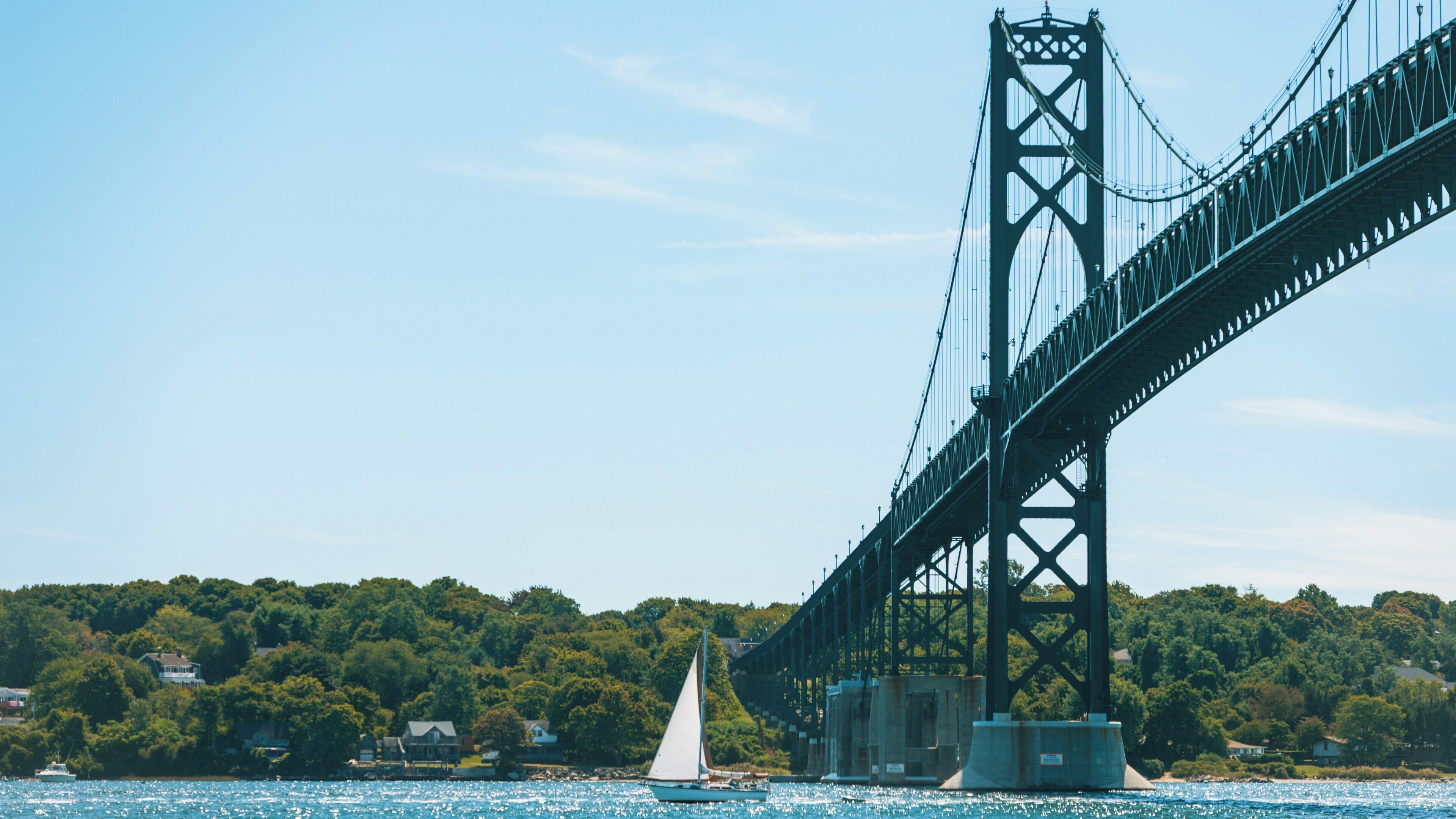 Newport Bridge spans the waters of Portsmouth, Rhode Island with a sailboat gliding beneath in bright sunshine