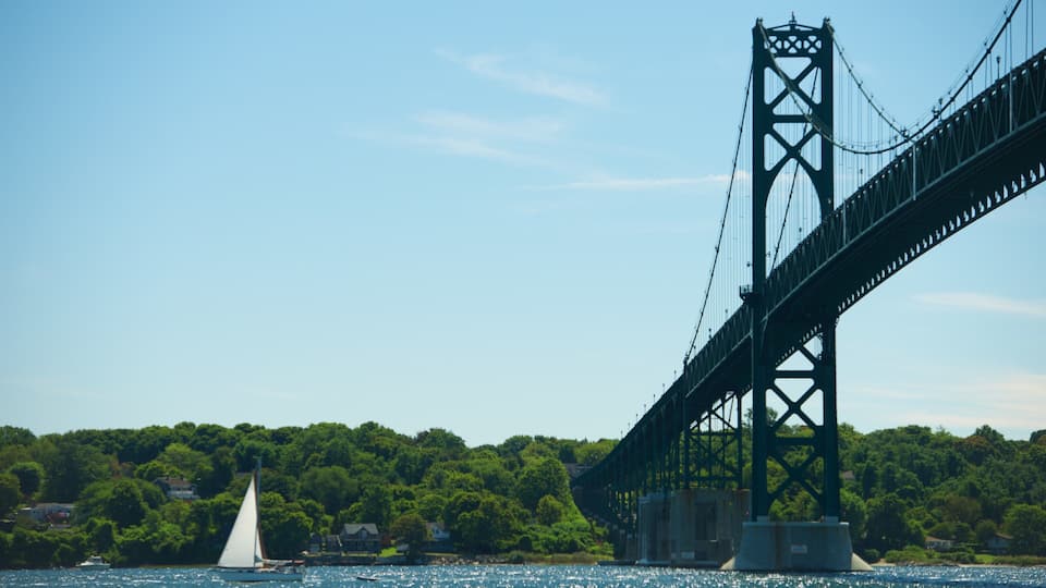 A large suspension bridge over a body of water with sailboats and a forested hillside in the background.