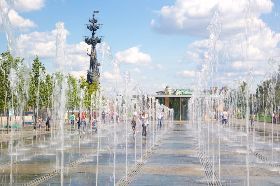 A water fountain plaza with multiple water jets, a central statue, and a distant building.