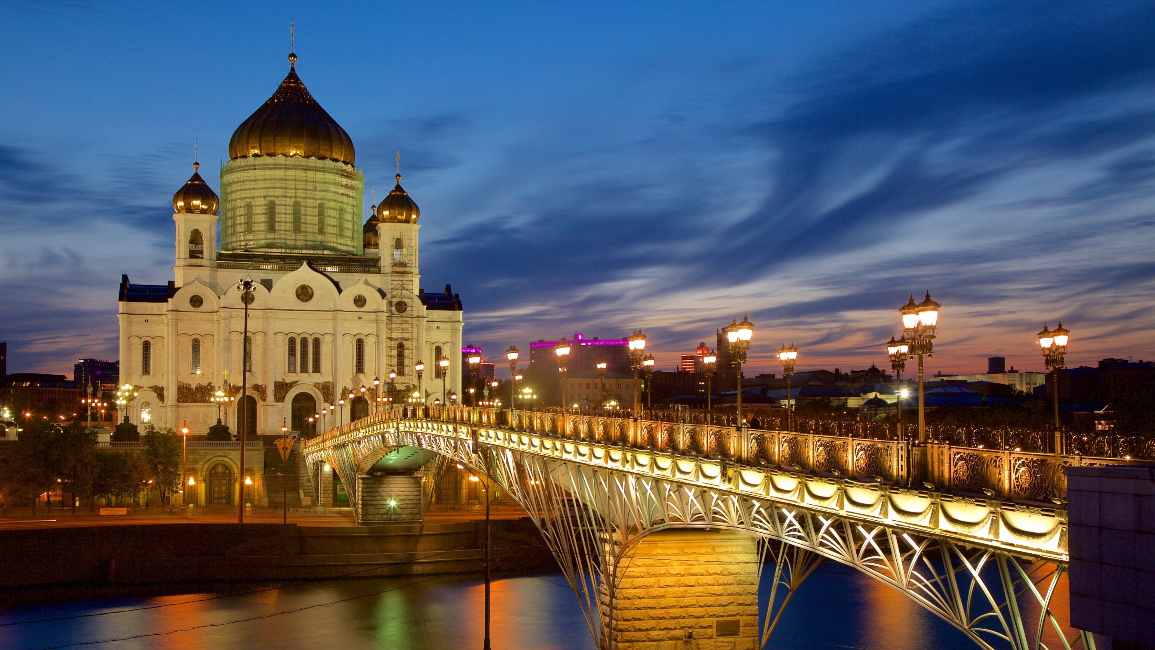 A bridge with ornate metalwork spanning a river, leading to a large white building with a golden dome.