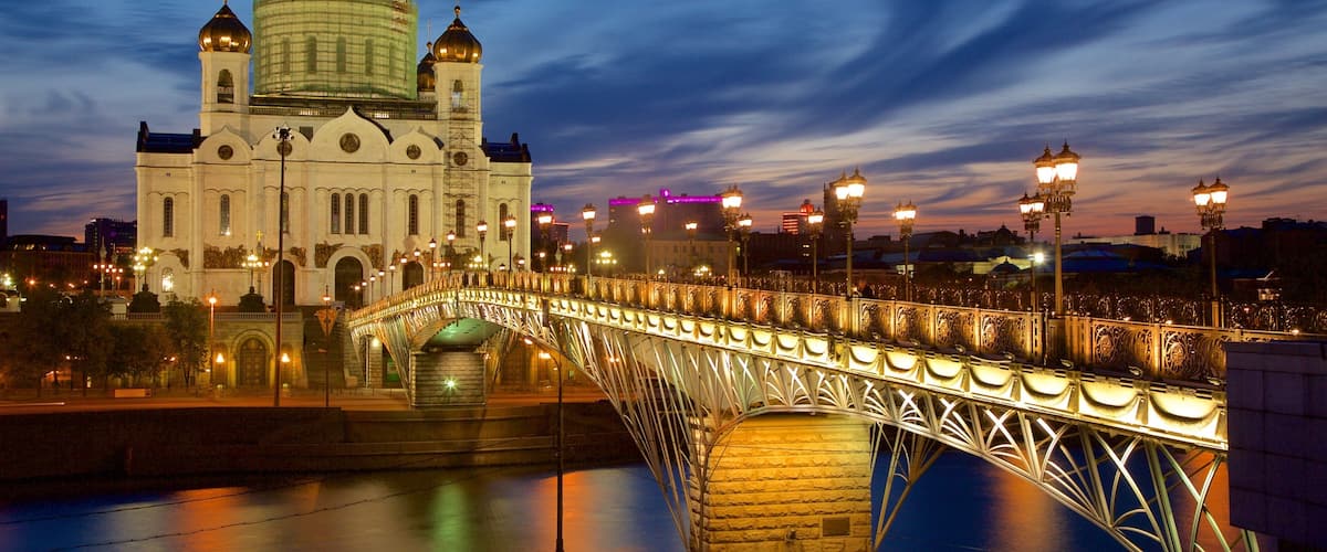 A bridge with ornate metalwork spanning a river, leading to a large white building with a golden dome.