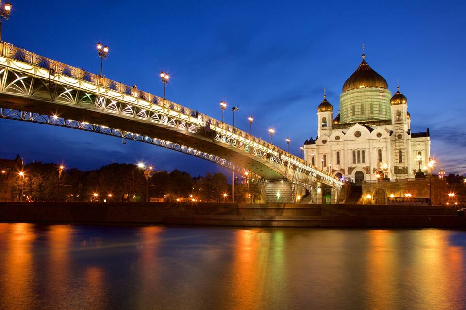 Un ponte con ringhiere elaborate e luci che scavalca un fiume, con un grande edificio a cupola sullo sfondo.