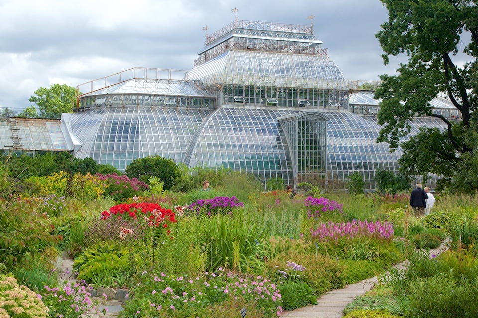 Ein großes Gewächshaus aus Glas, umgeben von einem vielfältigen Garten mit verschiedenen Blumen und Pflanzen.