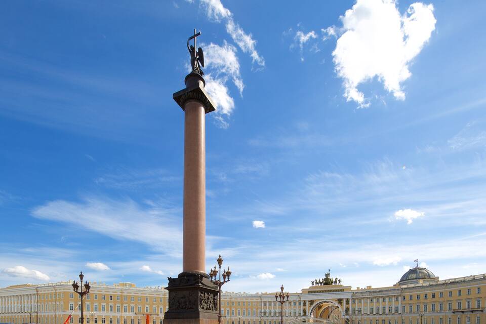 Un alto monumento con una statua in cima, sullo sfondo di un grande edificio giallo con una cupola e un cielo azzurro intenso.