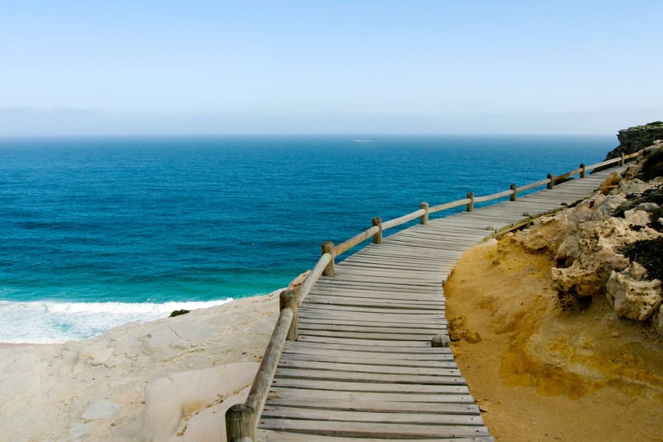 Ein hölzerner Steg führt an einer felsigen Küste entlang, im Hintergrund das Meer.