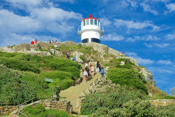 A lighthouse on a hill with a staircase leading up to it, surrounded by greenery and rocky terrain.
