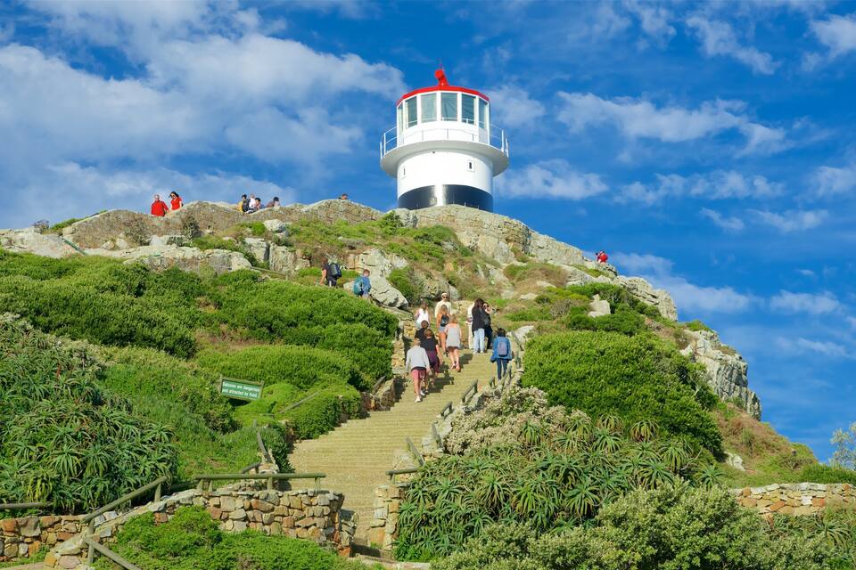 A lighthouse on a hill with a staircase leading up to it, surrounded by greenery and rocky terrain.