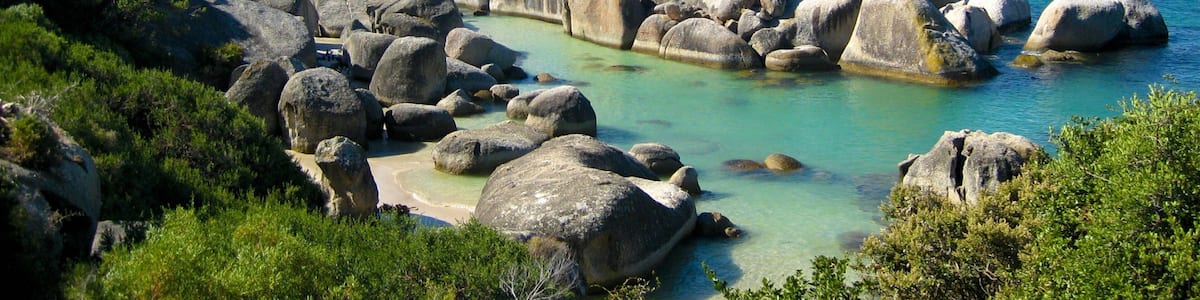 A coastal landscape with large boulders, clear turquoise waters, and distant mountains.