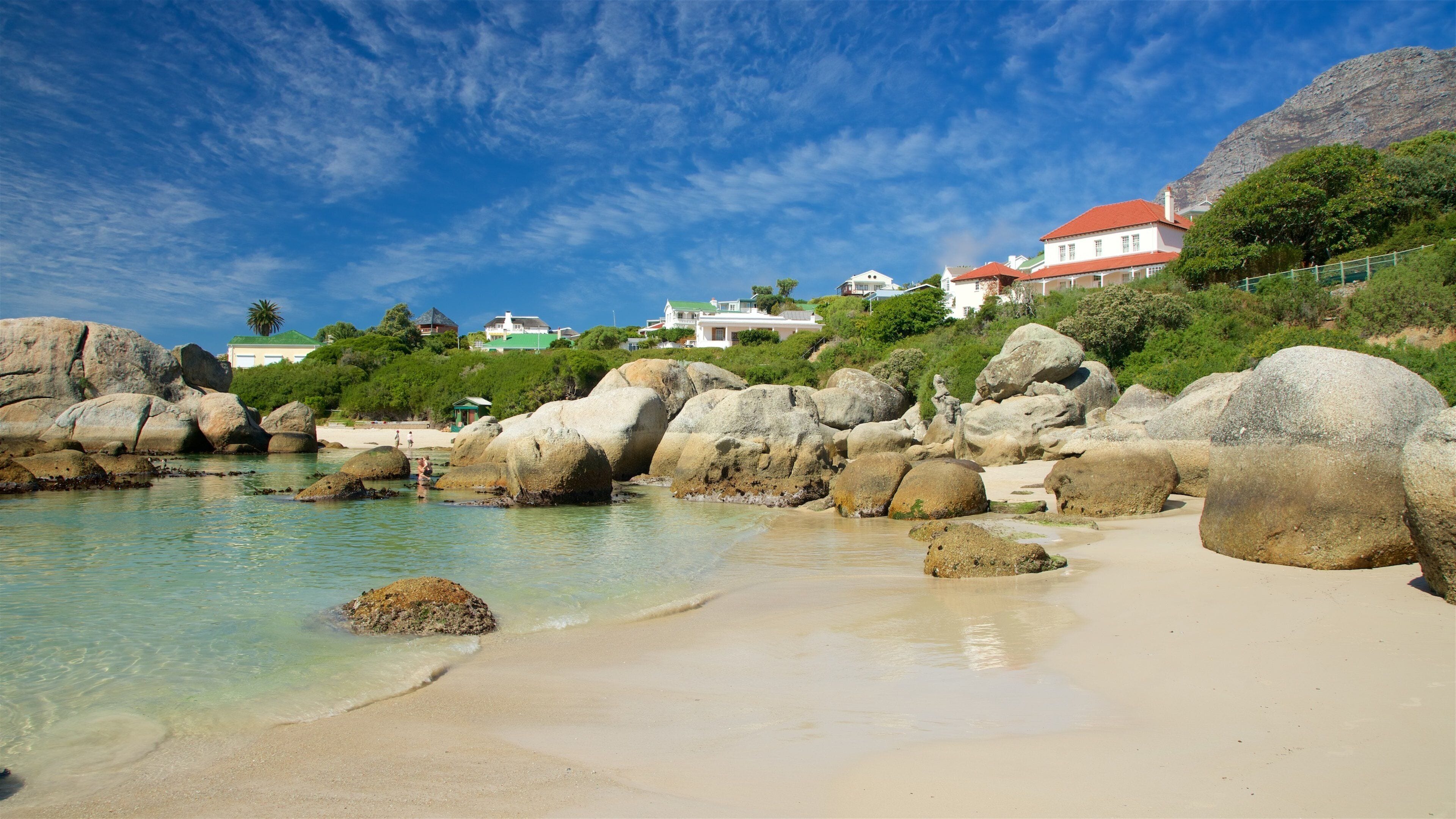 A coastal beach with large boulders, clear turquoise water, and houses with red roofs in the background.