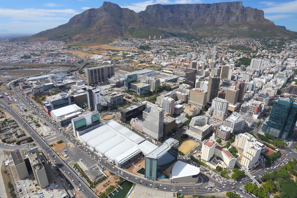 A cityscape with modern buildings, a large stadium, and mountains in the background.
