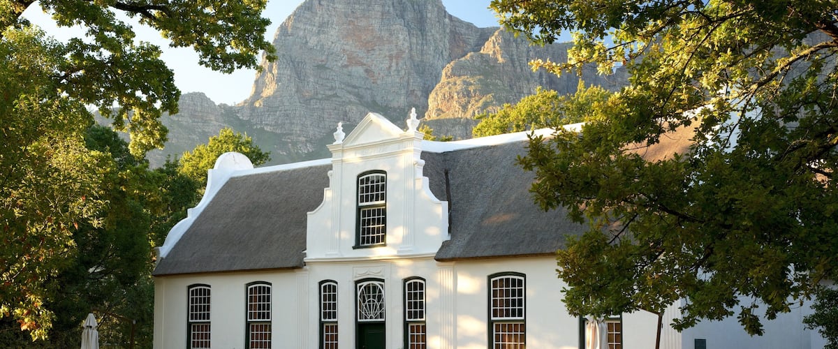 A white colonial-style building with a steeply pitched roof, surrounded by lush greenery and a mountainous backdrop.