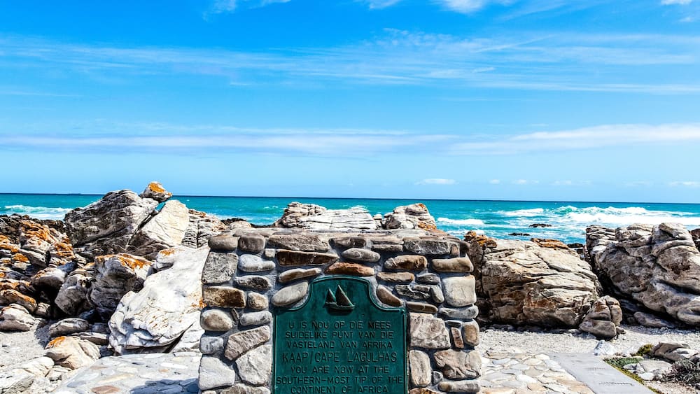Um monumento de pedra com uma placa, um trecho de costa rochosa e os oceanos Índico e Atlântico.