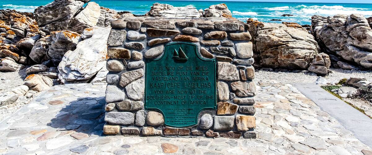 A stone monument with a plaque, a rocky shoreline, and the Indian Ocean and Atlantic Ocean.