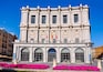Un bâtiment de style classique, doté d’arches et de colonnes, entouré de fleurs roses et surplombé d’un ciel bleu clair.