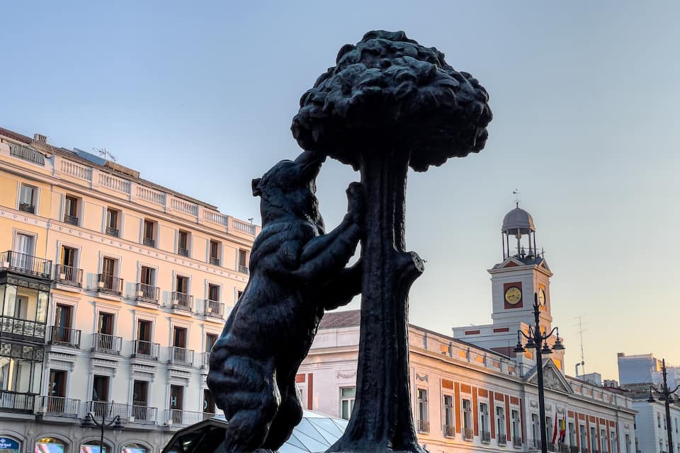 A statue of a bear reaching up to a tree, with a clock tower and buildings in the background.